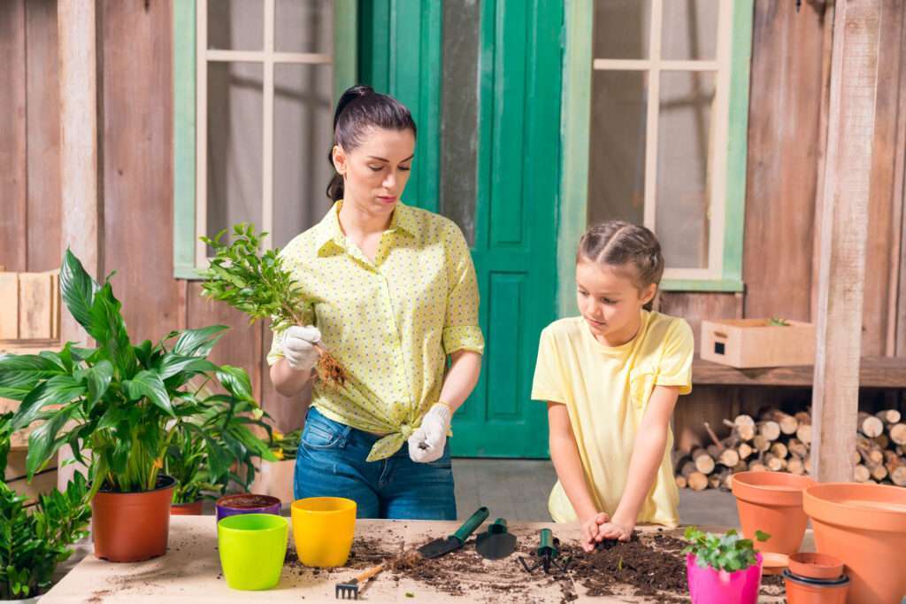 A woman and a young girl are engaged in gardening activities, with the woman holding a plant and the girl using a trowel on soil, surrounded by colorful pots and gardening tools.