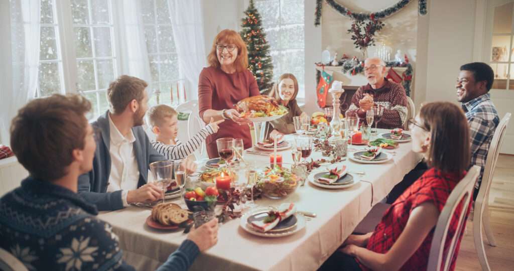 A group of people gathered around a dining table set for a festive meal. A woman is serving a roast turkey. The room is decorated for the holidays with a Christmas tree and garlands. Various dishes and drinks are on the table.