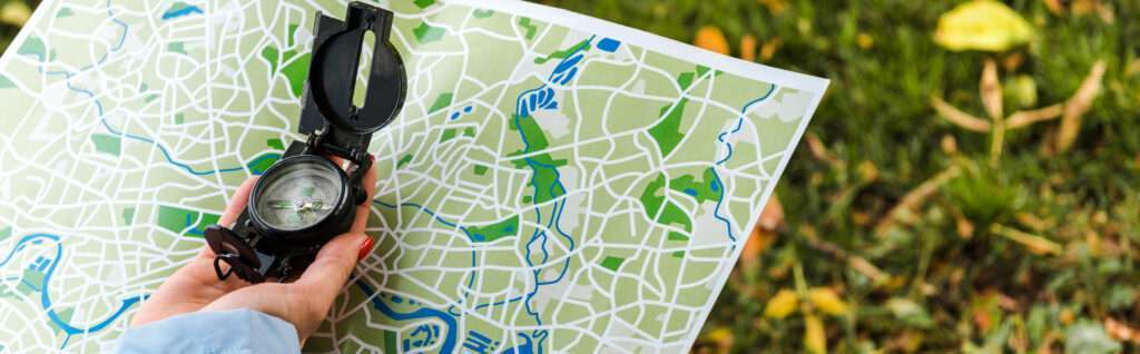 A person holds a compass in their left hand while looking at a detailed city map. The map displays various streets and green areas, suggesting parks. The background shows grass and scattered leaves, indicating an outdoor setting.