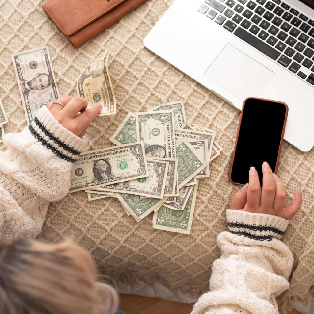 A person wearing a knitted sweater is counting U.S. dollar bills on a patterned surface. Nearby are a smartphone, a laptop, and a brown wallet. The phone screen is blank.